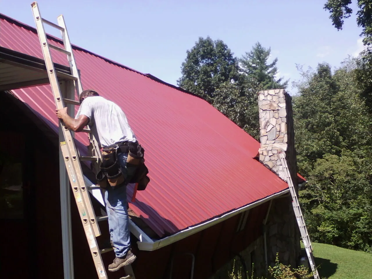 Roofer on a ladder beside a red metal roof and stone chimney — Soffit & Fascia Repair by Roofing Brothers in North Berwick